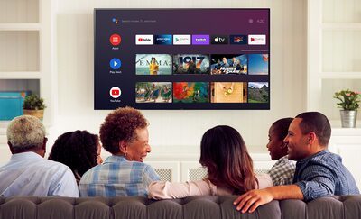 Familie și prieteni stând pe canapea, cu meniul de divertisment Android TV pe un televizor montat pe perete