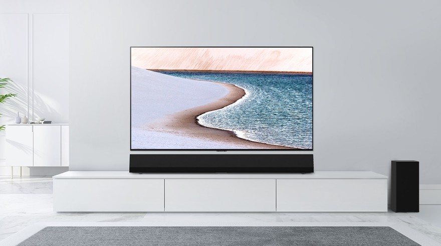 Un televizor amplasat pe un perete gri deschis. Bara de sunet LG se află dedesubt, pe un dulap alb. Televizorul afișează o plajă.