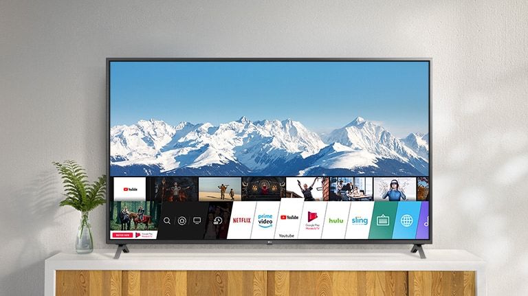 Televizor amplasat pe un suport alb, pe un perete alb. Ecranul TV prezintă ecranul principal cu webOS.