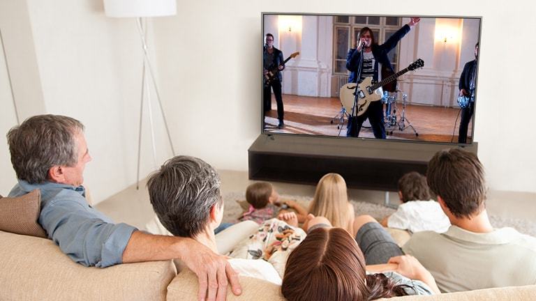 O familie cu șapte membri stau în camera de zi și se uită la un film. Pe ecranul TV apare o formație care cântă.